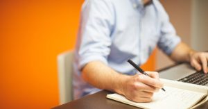 man writing at desk with laptop
