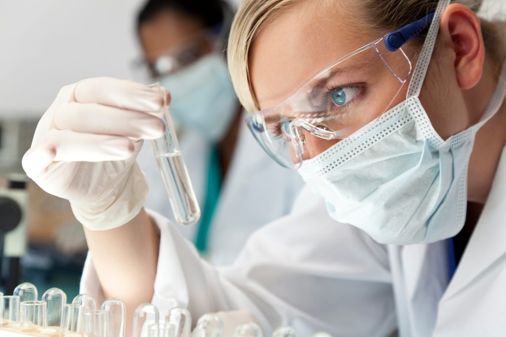 Blonde woman in a lab setting looking at a clear tube while wearing protective glasses, gloves and a face mask